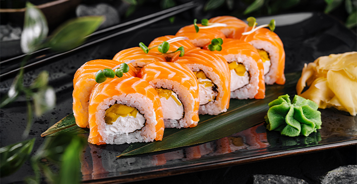 Sushi rolls topped with salmon, garnished with microgreens, served on a dark plate with wasabi and ginger.