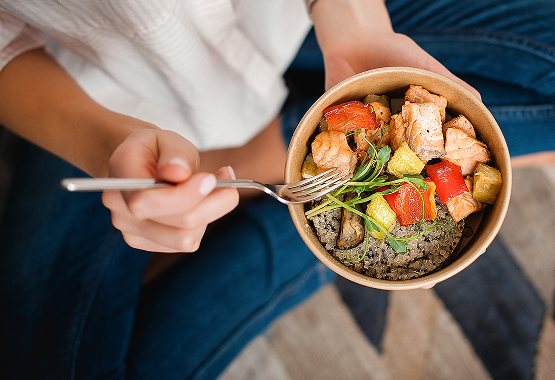 Person holds a bowl of salmon, vegetables, and grains, using a fork. Wearing jeans, seated on a patterned surface.
